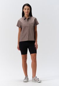 Lichtbruin overhemd met korte mouwen en een borstzak, gecombineerd met zwarte shorts en grijze sandalen, tegen een effen achtergrond.