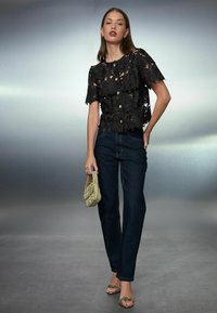 Sort blonderbluse med blomsterprint, korte ærmer og guldknapper, kombineret med mørk denimjeans og en perlebesat guldtaske.