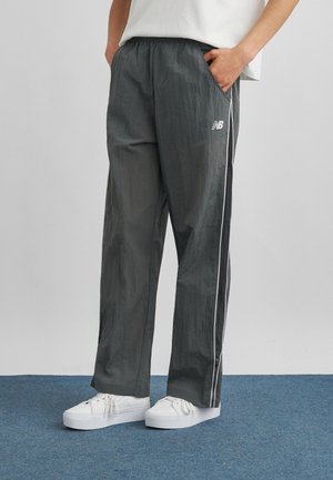 Person iført løse sorte New Balance træningsbukser med hvide sidelommer og hvide sneakers, stående på blåt tæppe.