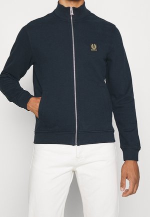Uomo che indossa una giacca nera con zip e un piccolo logo sul petto, pantaloni bianchi, una mano in tasca, su sfondo neutro.