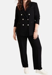 Zwarte dubbelgeknoopte blazer met witte knopen en contrasterende stiksels, gecombineerd met zwarte broek en witte sneakers.