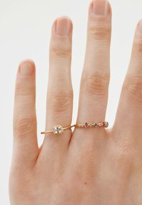 Anillos de oro en una mano, uno con una gema clara en un engaste cuadrado y el otro con piedras de colores en un diseño lineal. Textura suave.