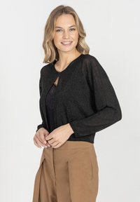 Czarny, metaliczny sweter z dzianiny o zaokrąglonym dekolcie, długich rękawach i pojedynczym zapięciu na guzik. Noszony na czarną bluzkę i beżowe spodnie.