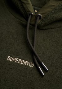 Sudadera con capucha de forro polar verde oliva, con cordones ajustables negros, que presenta un logo bordado de "SUPERDRY" en color crema, textura suave y diseño con capucha.