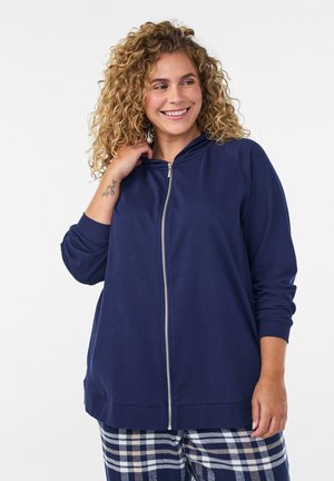 Krullendharige vrouw die lacht, draagt een navy zip-up hoodie en geruite broek, met een zichtbare tatoeage op haar pols tegen een effen achtergrond.