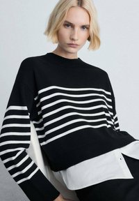 Schwarz-weißer gestreifter Pullover mit Rundhalsausschnitt, gerippten Bündchen und Saum. Darüber trägt er ein weißes Hemd mit Kragen, das ein strukturiertes Strickmuster zeigt.