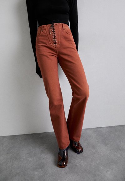 Roestkleurige high-waisted jeans met een voorzijde vetersluiting, rechte pijpen en een contrasterende zwarte top. Gemaakt met een patroon van hoge laarzen.