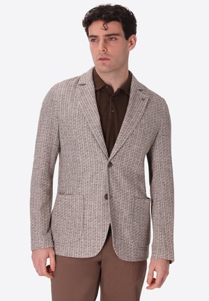 Uomo che indossa un blazer scozzese marrone chiaro sopra una camicia marrone scuro con colletto e pantaloni marroni coordinati, che guarda leggermente di lato.