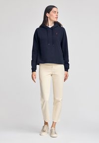 Felpa blu navy con tasca frontale e cappuccio con coulisse, abbinata a pantaloni slim-fit color crema e sneakers chiare. Design semplice e texture morbida.