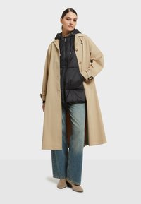 Beige trenchcoat med knyts midja över en svart pufferväst, parat med ljusblå jeans och beige ankelstövlar. Släta tyger och avslappnad passform.