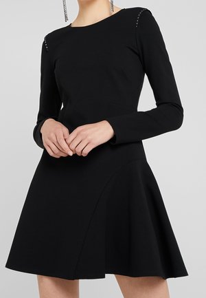 Kobieta ubrana w czarną sukienkę z długim rękawem, rozszerzaną na dole, ozdobioną małymi srebrnymi ćwiekami na szwach ramion oraz z wiszącymi kolczykami z łańcuszków.