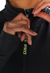 Haut de sport noir avec fermeture éclair et col montant. Présente un logo "Nike Pro" vert et une texture lisse. Mains en train d'ajuster la fermeture éclair.