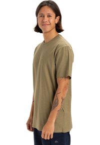 Joven con cabello oscuro de longitud media, vistiendo una camiseta verde oliva y pantalones azul marino, sonriente y de pie de lado con tatuajes visibles en los brazos.