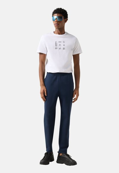 Młody mężczyzna w za dużych niebieskich okularach przeciwsłonecznych, białej koszulce z czarnymi ikonami, granatowych spodniach i czarnych tenisówkach, stojący na białym tle.