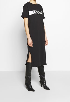 Femme portant une robe midi noire avec un logo blanc imprimé sur la poitrine et des bottes noires à talons hauts jusqu'aux genoux, debout sur un sol clair.