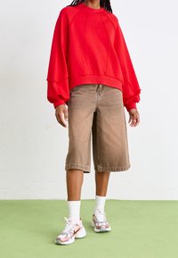 Red sweatshirt with oversized sleeves and ribbed hem, paired with brown denim shorts that extend to the knee. White socks and multicolored sneakers.