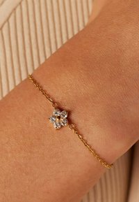 Gouden kettingarmband met een bloemontwerp, waarbij heldere edelstenen als bloemblaadjes dienen en een gouden centrum, rustend op een gestructureerd huidoppervlak.