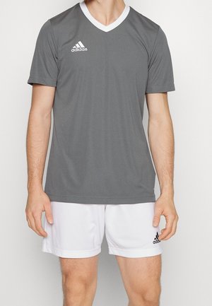 Homme portant un t-shirt gris Adidas à manches courtes et col en V et un short blanc Adidas, debout devant un fond uni.