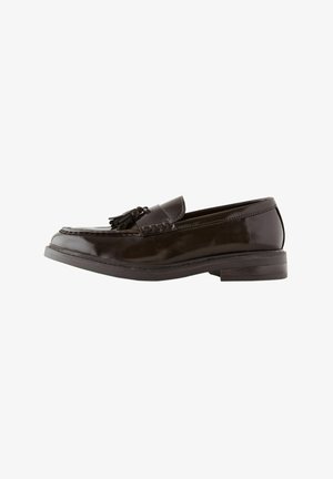 Mocasín de cuero marrón con un acabado brillante, que presenta un adorno de borlas anudadas y costuras decorativas a lo largo del borde. Tacón bajo y plano.