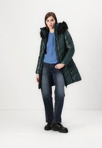 Cappotto imbottito verde scuro con cappuccio bordato di pelliccia, chiusura con zip e tasche laterali. Indossato sopra un maglione blu, abbinato a jeans scuri e scarpe nere.