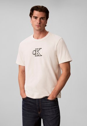 Mann trägt ein off-white kurzärmeliges T-Shirt mit schwarzem "CK" Logo und dunkelblaue Jeans, steht mit Händen in den Taschen vor einem einfarbigen Hintergrund.