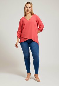 Top corallo con scollo a v e maniche tre quarti, design sovrapposto, abbinato a jeans skinny blu e sandali metallici. Tessuto dalla texture liscia.