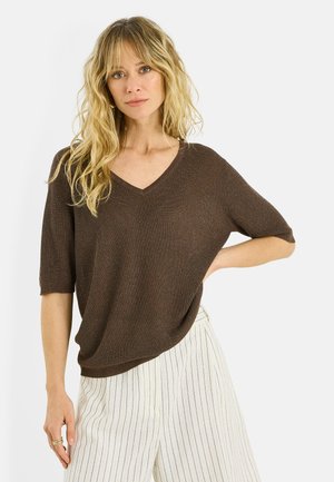 Mujer con cabello rubio largo, vestida con un top marrón de punto con escote en V y pantalones blancos con delgadas rayas negras verticales, posando con una mano en la cadera.