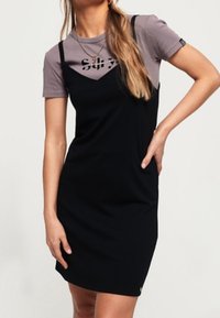 Svart ärmlös klänning med tunna axelband, bärs över en grå t-shirt med tryckt logotyp. Slät textur och figurnära design.