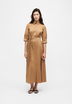 Femme debout portant une longue robe beige boutonnée avec des manches trois-quarts, une ceinture à nouer et des sandales noires à bouts ouverts sur un fond uni.