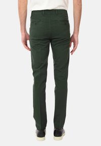 Chino in cotone verde con vestibilità dritta, dotati di due tasche posteriori con bottoni e cuciture decorative sui lati. Indossati con scarpe nere.
