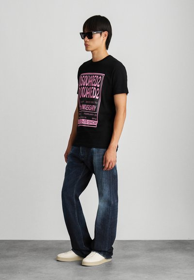 Camiseta negra con estampado gráfico rosa, vaqueros de denim azul y zapatillas color crema. Modelo con gafas de sol negras, de pie contra un fondo neutro.