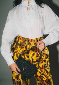 Borsa nera con perline e nappina, indossata con una camicetta bianca dal colletto alto e pantaloni gialli a motivi floreali con una cintura multicolore.