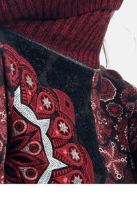 Col roulé rouge à motifs avec une texture en gros tricot, présentant des dessins graphiques complexes en noir et rouge, soulignés par des accents argentés.