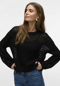 Zwarte gebreide trui met een losse pasvorm, voorzien van een gestructureerd, opengeweven ontwerp en lange mouwen. Gemaakt in combinatie met blauwe jeans.