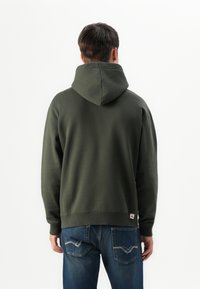 Man seen from the back wearing a dark green hooded sweatshirt and blue jeans with visible pocket stitching on a plain white background.