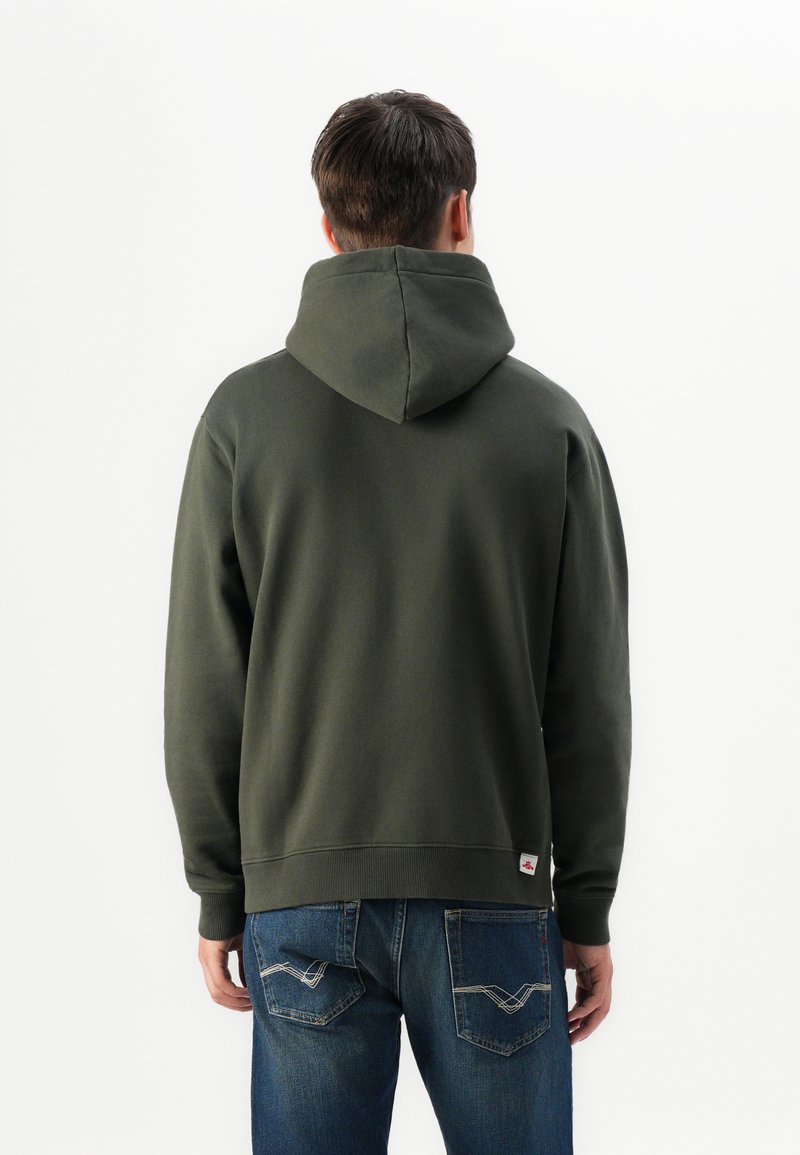 Uomo visto da dietro che indossa un felpa con cappuccio verde scuro e jeans blu con cuciture sulla tasca visibili, su uno sfondo bianco semplice.