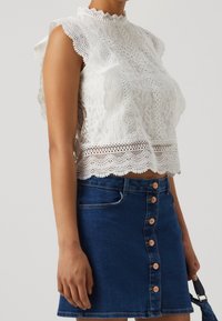 Top corto in pizzo bianco con collo alto e bordi ondulati, abbinato a una gonna di jeans blu con chiusura frontale a bottoni e tasche.