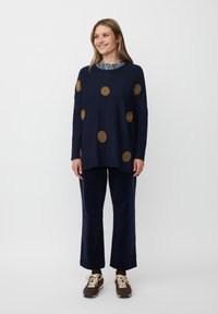 Granatowy sweter z dużymi złotymi okręgami, zestawiony z granatowymi spodniami o szerokich nogawkach i brązowymi sneakersami, z wzorzystym kołnierzykiem widocznym pod spodem.