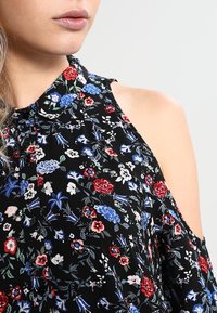 Blusa nera floreale con colletto, chiusura a bottoni e design a spalla scoperta. Presenta vari motivi floreali in rosso, blu e bianco.