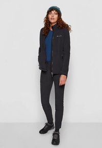 Φλις μπουφάν σε μαύρο χρώμα με ψηλό γιακά, κλείσιμο με φερμουάρ, φορεμένο πάνω από μπλε μπλούζα και μαύρο παντελόνι. Υφή υφάσματος με ανάγλυφα μοτίβα; συνδυασμένο με σκούρα υποδήματα.