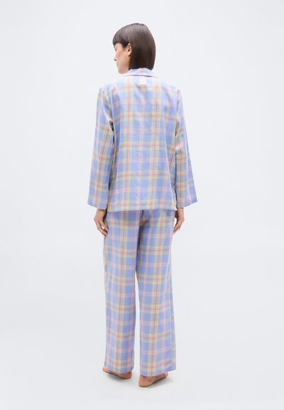 Γυναίκα όρθια, ξυπόλυτη, γυρισμένη πλάτη, φοράει αέρινο πιτζάμα σε παστέλ καρό με μακριά μανίκια και φαρδιά παντελόνια, μπροστά σε απλό φόντο.