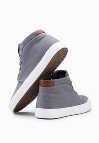 Zapatillas altas de lona gris con suelas de goma blancas. Presenta un acento de cuero marrón en el talón y cordones con seis ojetes.