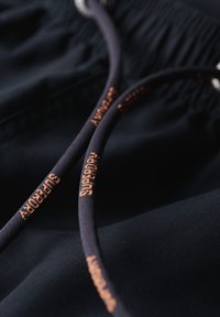 Shorts de tela de color navy con cinturilla elástica y dos cordones. Uno de los cordones presenta "Superdry" en costura naranja. Textura suave.