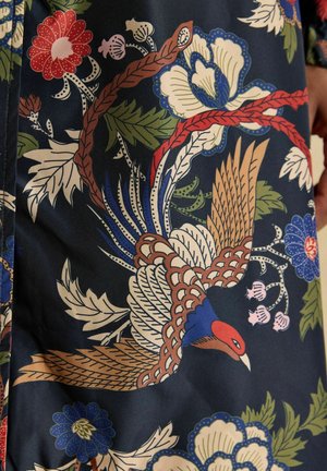 Buntes Stoffmuster mit einem braunen, blauen und roten Vogel im Flug, umgeben von verschiedenen stilisierten Blumen und grünen Blättern auf dunklem Hintergrund.