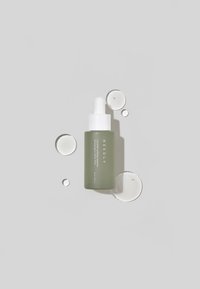 Bottiglia contagocce in vetro verde con tappo bianco, etichettata "NEEDLY", accanto a gocce di liquido trasparente su uno sfondo grigio.
