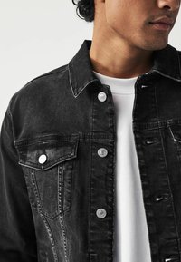 Schwarze Denimjacke mit silbernen Druckknöpfen, zwei Brusttaschen und einer lockeren Passform, über einem weißen T-Shirt getragen. Der Stoff hat eine abgenutzte und glatte Textur.