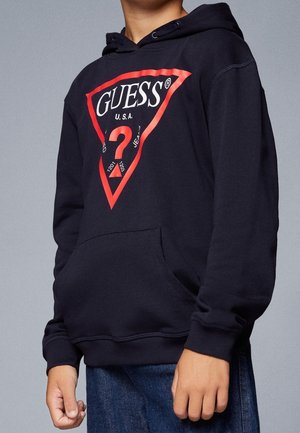 Person iført sort GUESS-hættetrøje med rødt trekantslogo og spørgsmålstegn, kombineret med blå jeans, stående mod en grå baggrund.