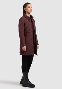 Abrigo burdeos acolchado con cuello alto, costura diagonal, dos bolsillos laterales y cierre frontal de cremallera, combinado con leggings negros y botas.