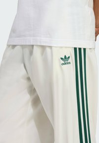 Jogging en élasthanne blancs avec trois bandes verticales vertes de chaque côté et un logo Adidas vert brodé sur la jambe gauche.