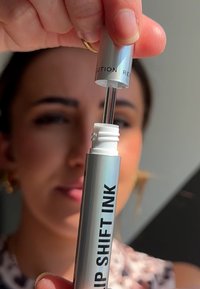 Sølv leppeprodukt tube merket "LIP SHIFT INK," med en klar applikator og hvit hette. Teksturert overflate synlig på tuben.